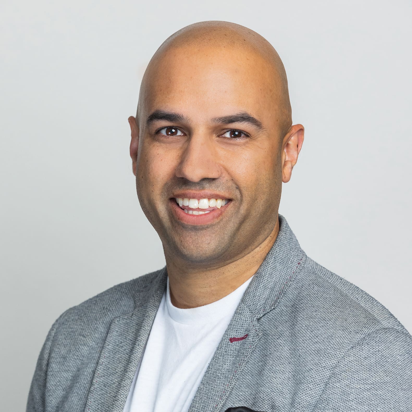 A smiling person with a bald head, wearing a grey blazer over a white shirt, against a light background.