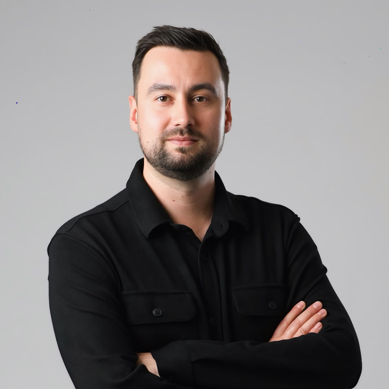 A man in a black shirt stands confidently with arms crossed against a gray background.