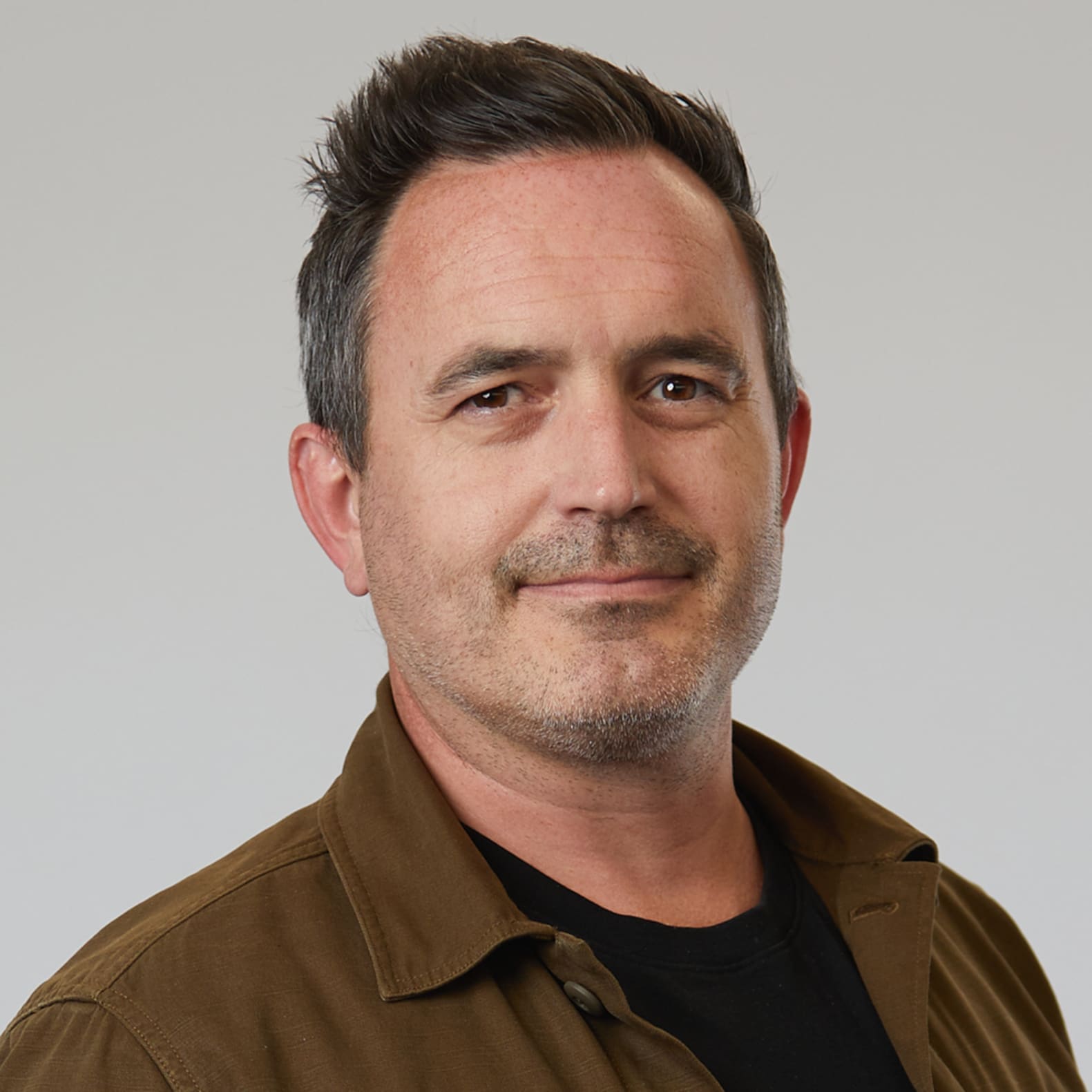 Man with brown hair and facial stubble wearing brown jacket over black shirt, smiling at camera