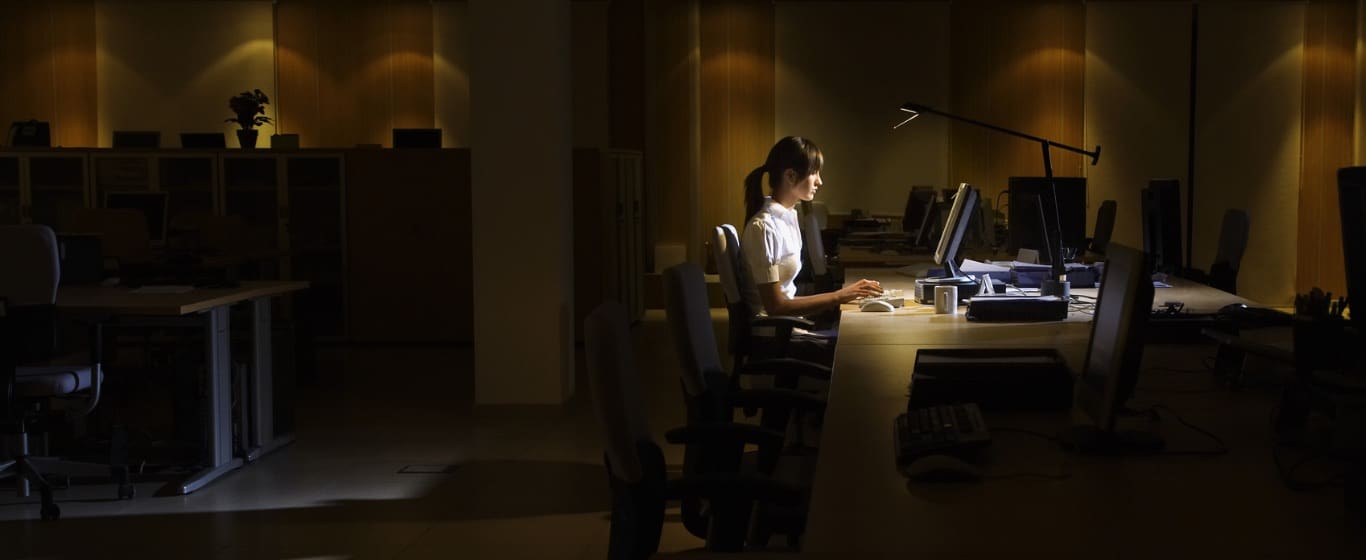 Person working alone at desk with computer in dimly lit office with desk lamp illumination