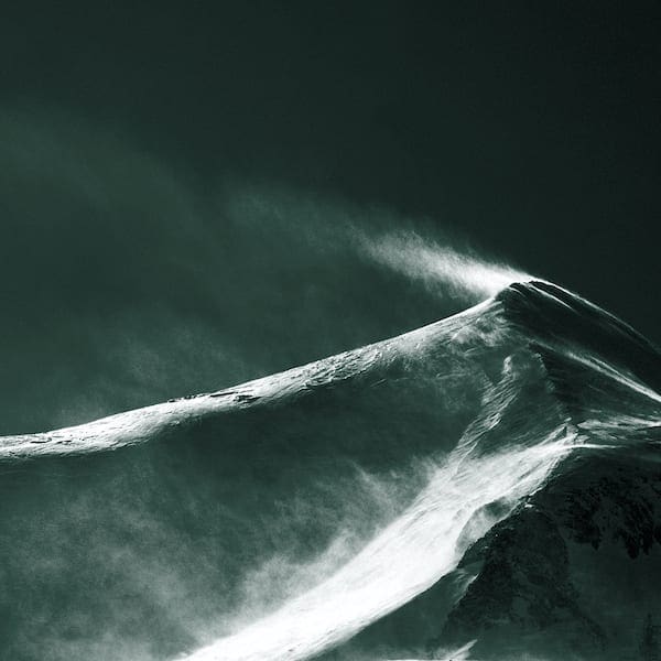 Snow-covered mountain peak with wind-blown snow against dark stormy sky