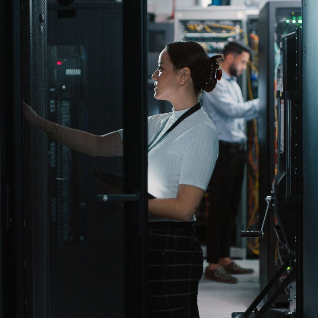 A woman checks a server rack while a man works in the background, surrounded by equipment.