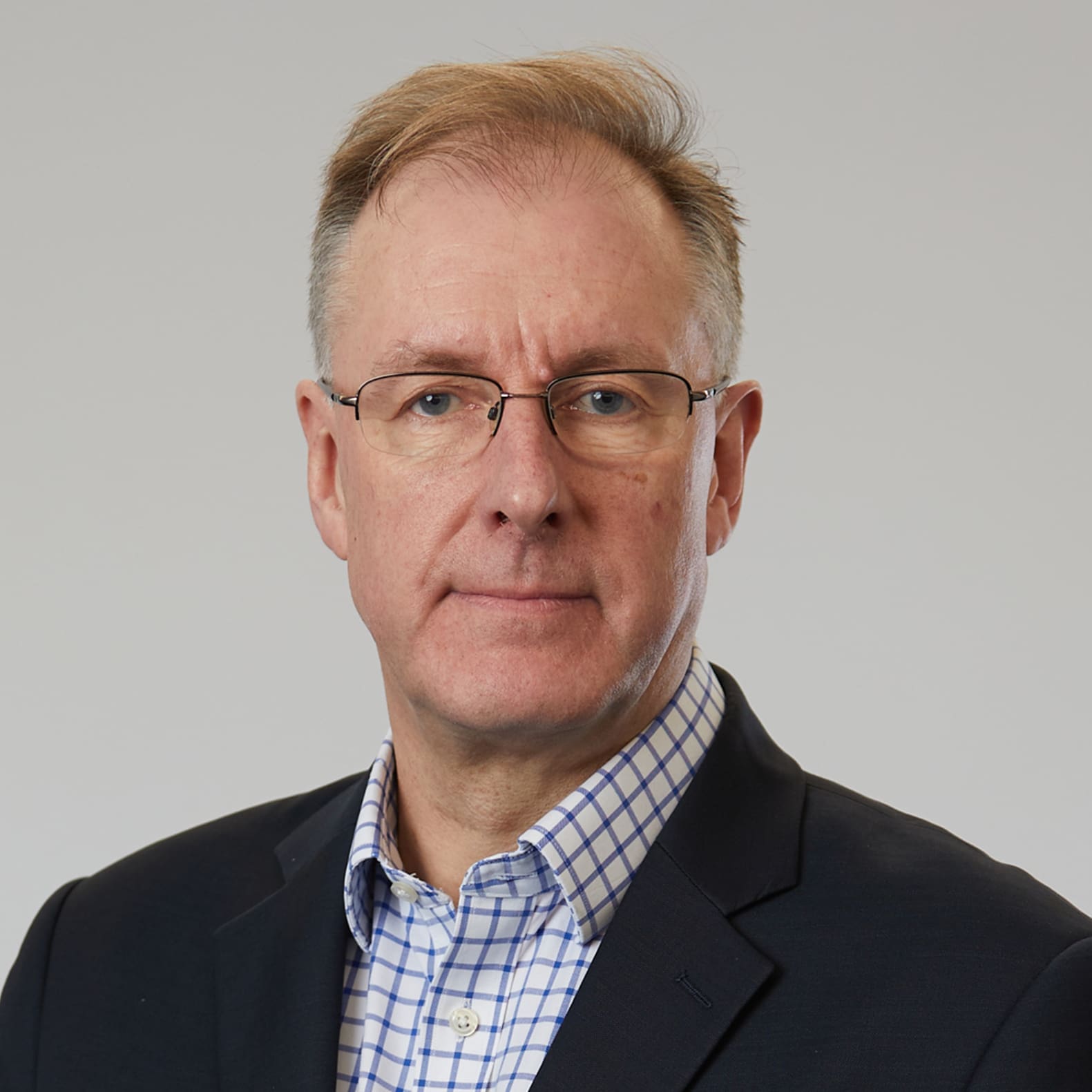 Professional headshot of middle-aged man in dark suit jacket, blue checkered shirt, and wire-rim glasses